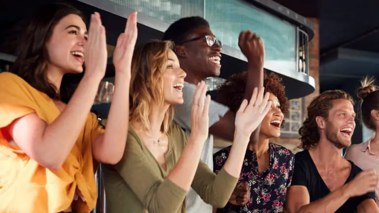 Group Of Male And Female Friends Celebrating Whilst Watching Game On Screen In Sports Bar